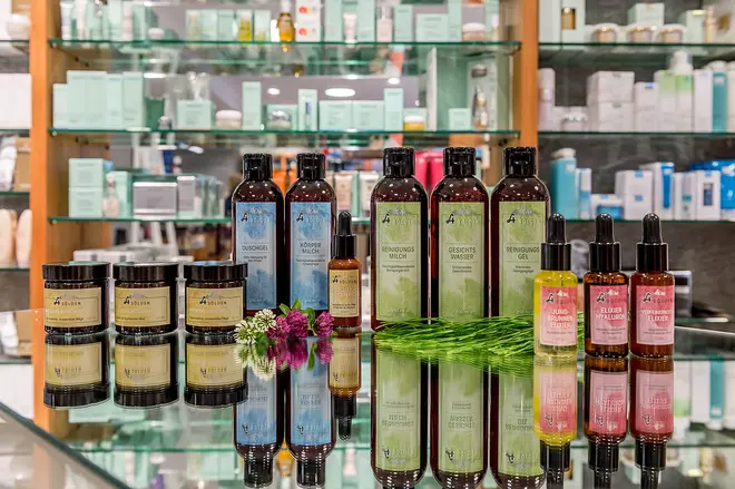 Verschiedene Flaschen mit Hautpflege- oder Schönheitsprodukten stehen fein säuberlich auf einer spiegelnden Glastheke, vor der kleine grüne Pflanzen und lila Blumen stehen, neben Regalen, die mit anderen Produkten in einer Apotheke gefüllt sind. Es handelt sich um Produkte der Naturkosmetik.