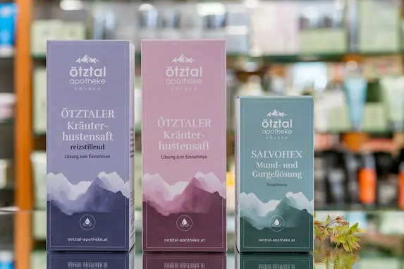 Auf einer Theke mit unscharfen Produktregalen im Hintergrund stehen drei Hautpflegeboxen mit minimalistischen Bergmotiven in Lila-, Rosa- und Grüntönen. Neben den Boxen steht eine kleine Pflanze. Es handelt sich um Produkte der Eigenherstellung.