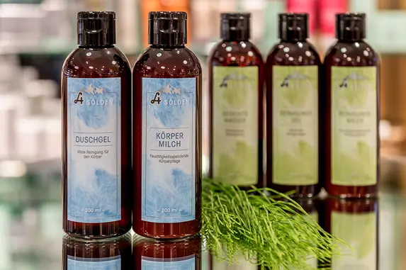 Fünf braune Flaschen mit blauen und grünen Etiketten sind auf einer spiegelnden Fläche ausgestellt, davor eine kleine grüne Pflanze. Der Hintergrund ist unscharf. Gezeigt werden Produkte der Naturkosmetik.