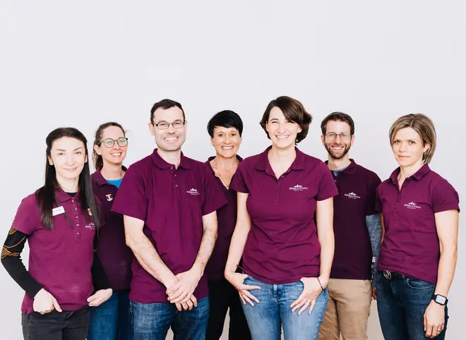 Sieben Personen stehen in einer Reihe vor einem schlichten weißen Hintergrund, alle tragen passende Oberteile und lächeln in die Kamera. Gezeigt wird das Team der Ötztal Apotheke.