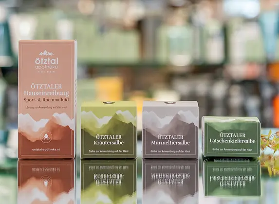 Eine Reihe bunter Verpackungen, die auf einer spiegelnden Oberfläche ausgestellt sind, mit Regalen mit verschwommenen Produkten im Hintergrund. Die bunten Verpackungen sind Produkte aus der Eigenherstellung der Apotheke Ötztal.