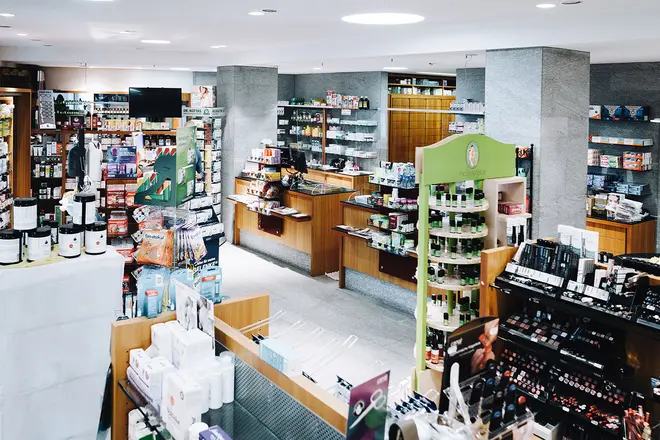 Ein hell erleuchtetes Ladeninnere mit Regalen, die mit verschiedenen Produkten bestückt sind, darunter Flaschen, Schachteln und Kosmetikartikel. Der Raum wirkt aufgeräumt, die Auslagen und Theken sind ordentlich in der ganzen Apotheke angeordnet.