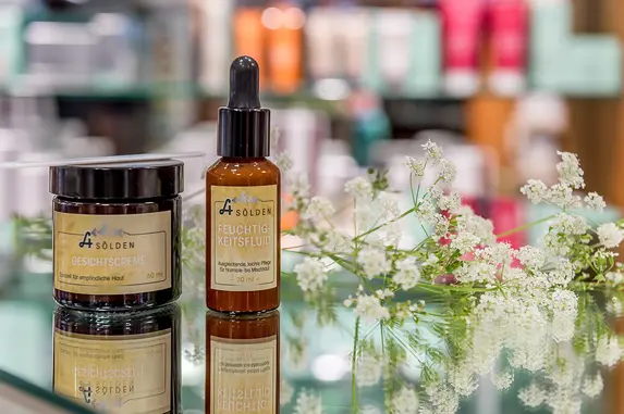 Zwei Hautpflegeprodukte in braunen Behältern stehen auf einer Glasfläche neben kleinen weißen Blumen, mit unscharfen Regalen im Hintergrund.