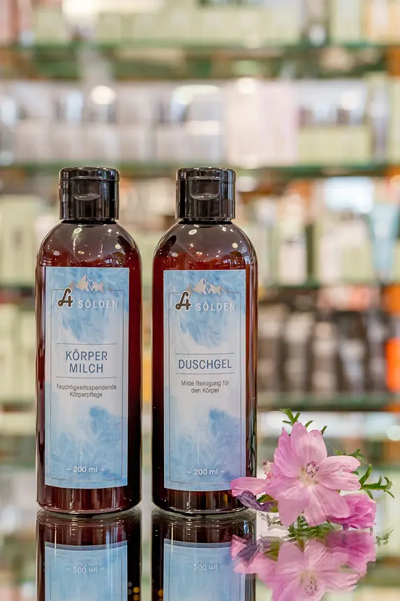 Zwei braune Flaschen mit blauen Etiketten sind nebeneinander auf einer Glasfläche ausgestellt, in der Nähe befindet sich eine rosa Blume. Im Hintergrund sind verschwommene Regale mit verschiedenen Produkten zu sehen.