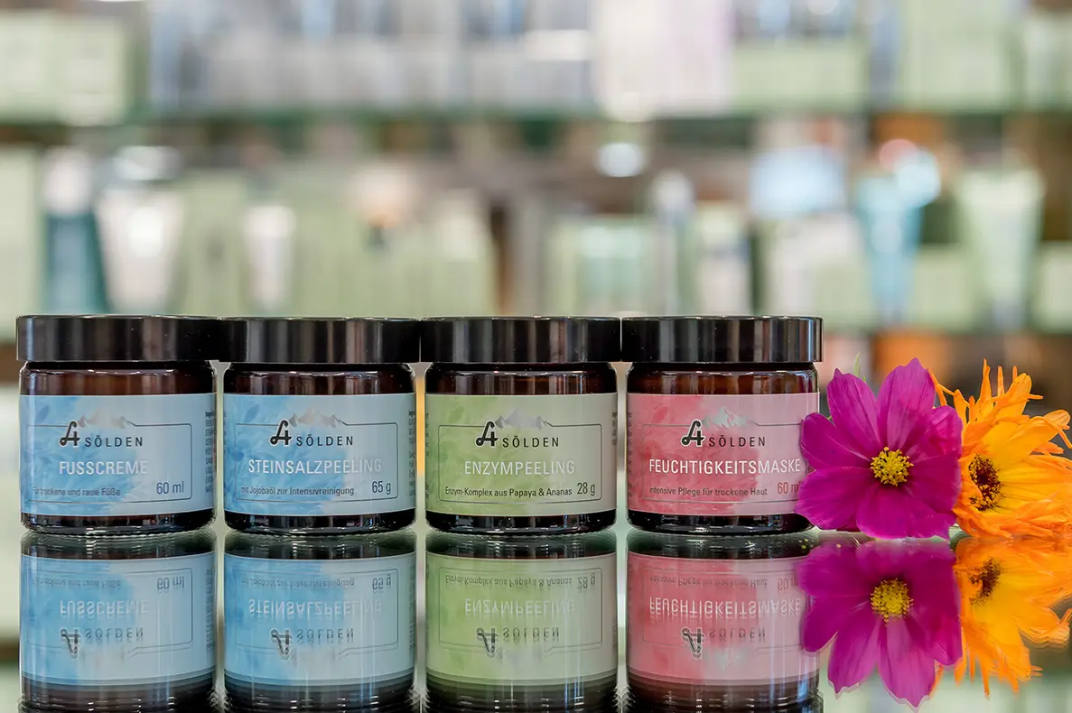 Vier Dosen mit Hautpflegeprodukten mit verschiedenfarbigen Etiketten sind in einer Reihe auf einer spiegelnden Oberfläche ausgestellt, daneben stehen zwei leuchtende Blumen - eine rosa und eine orange. Der Hintergrund ist unscharf.