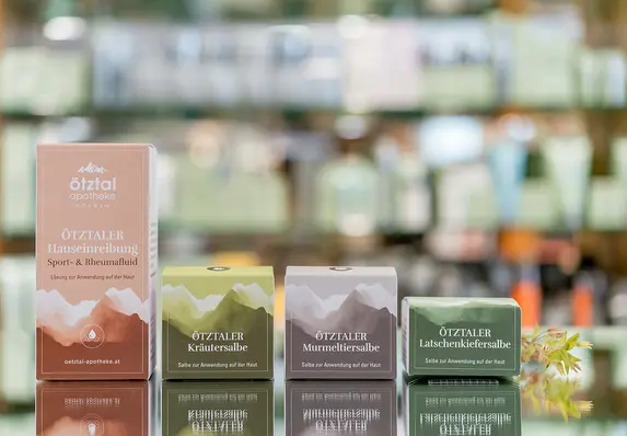 Eine Reihe bunter Verpackungen, die auf einer spiegelnden Oberfläche ausgestellt sind, mit Regalen mit verschwommenen Produkten im Hintergrund. Die bunten Verpackungen sind Produkte aus der Eigenherstellung der Apotheke Ötztal.