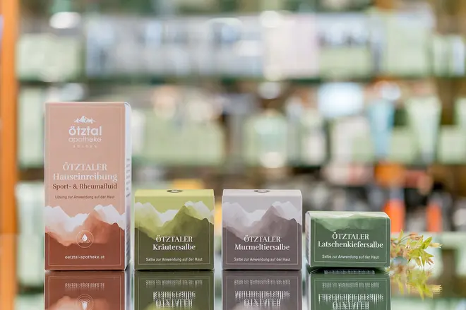 Eine Reihe bunter Verpackungen, die auf einer spiegelnden Oberfläche ausgestellt sind, mit Regalen mit verschwommenen Produkten im Hintergrund. Die bunten Verpackungen sind Produkte aus der Eigenherstellung der Apotheke Ötztal.
