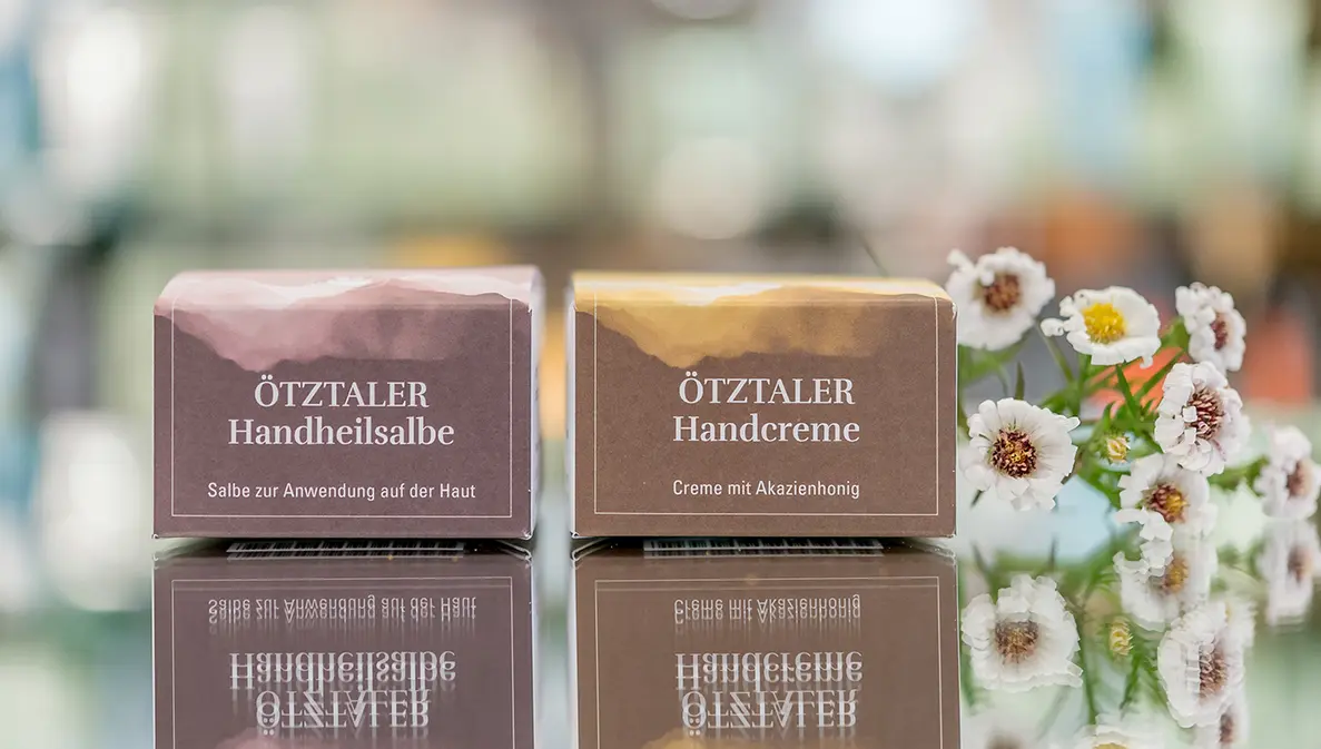 Zwei rechteckige Verpackungen in Braun- und Goldtönen liegen auf einer spiegelnden Oberfläche neben einem Strauß weißer Gänseblümchen vor einem weichen, unscharfen Hintergrund.